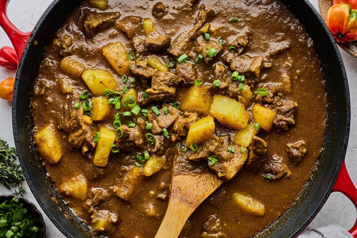 Curry goat in a large dutch oven with potatoes and garnished with parsley, with a wooden spoon stirring it