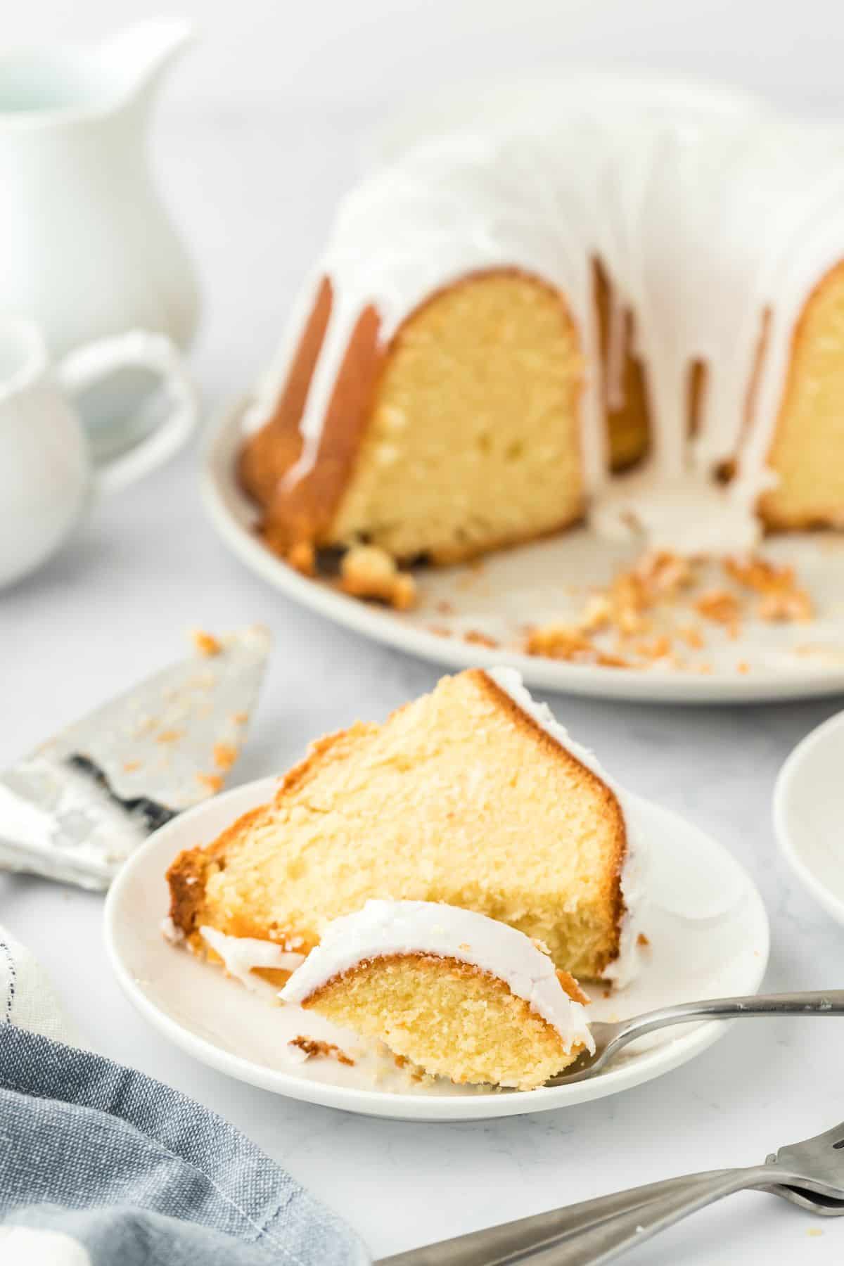 A slice of million dollar pound cake with white glaze on a white plate, with a fork resting beside it. In the background there's the rest of the cake on a serving platter