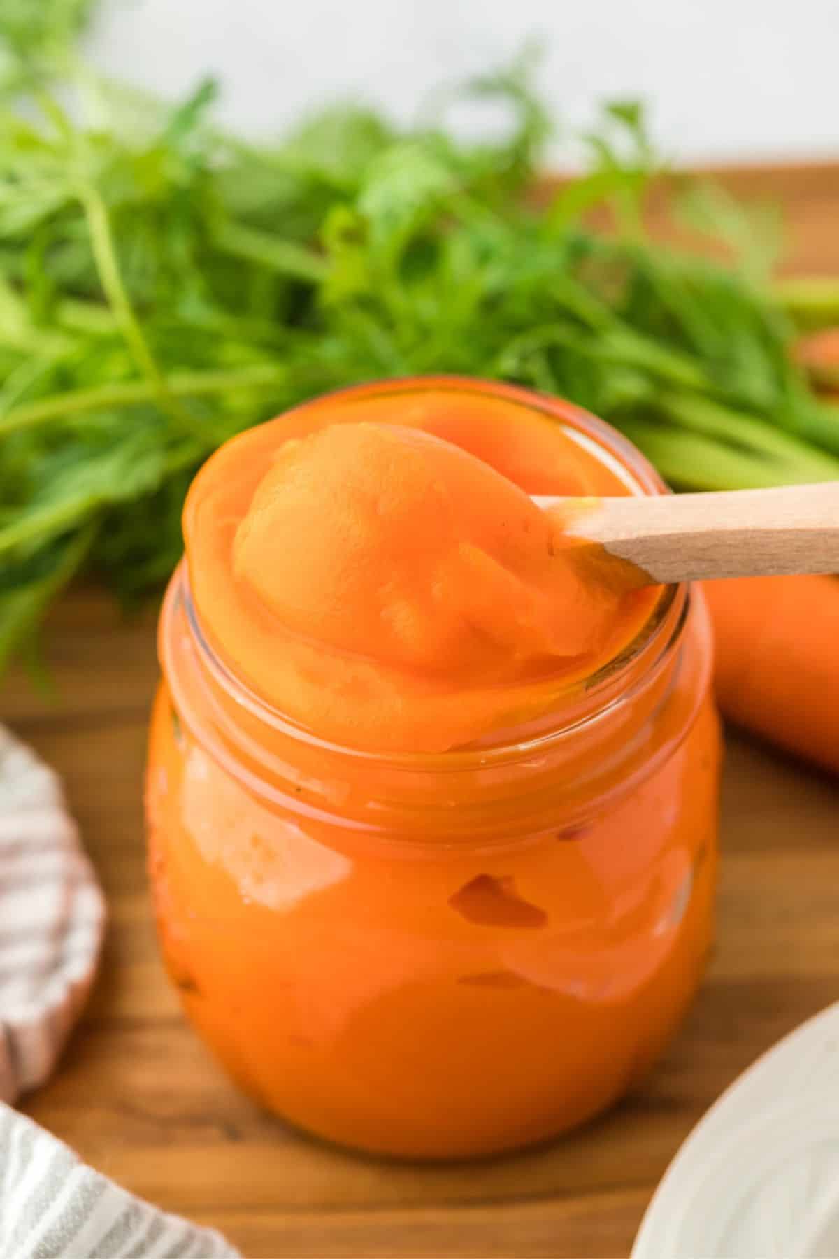 Closeup of a jar filled with smooth carrot puree being scooped with a wooden spoon, sitting in a wooden table with carrots in the background