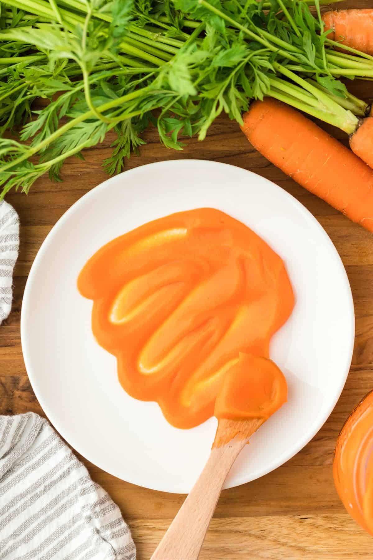 Overhead shot of a white plate with a swirl of carrot puree on a wooden table, a wooden spoon with puree on it, fresh whole carrots with green tops, and a striped kitchen towel