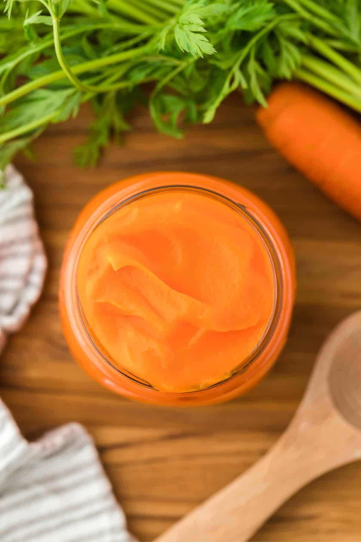 Overhead shot of a jar of carrot puree on a wooden table, with fresh carrots and their green tops and a wooden spoon next to it