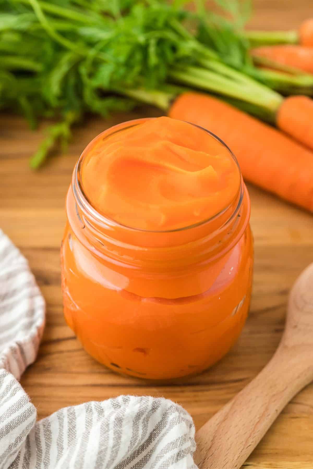 A jar of smooth carrot puree on a wooden table, with fresh carrots and their green tops and a wooden spoon next to it