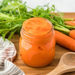 A jar of carrot puree on a wooden table, with fresh carrots and their green tops in the background, alongside a striped kitchen towel and a wooden spoon