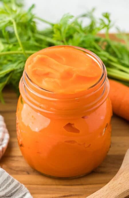 A jar of carrot puree on a wooden table, with fresh carrots and their green tops in the background, alongside a striped kitchen towel and a wooden spoon