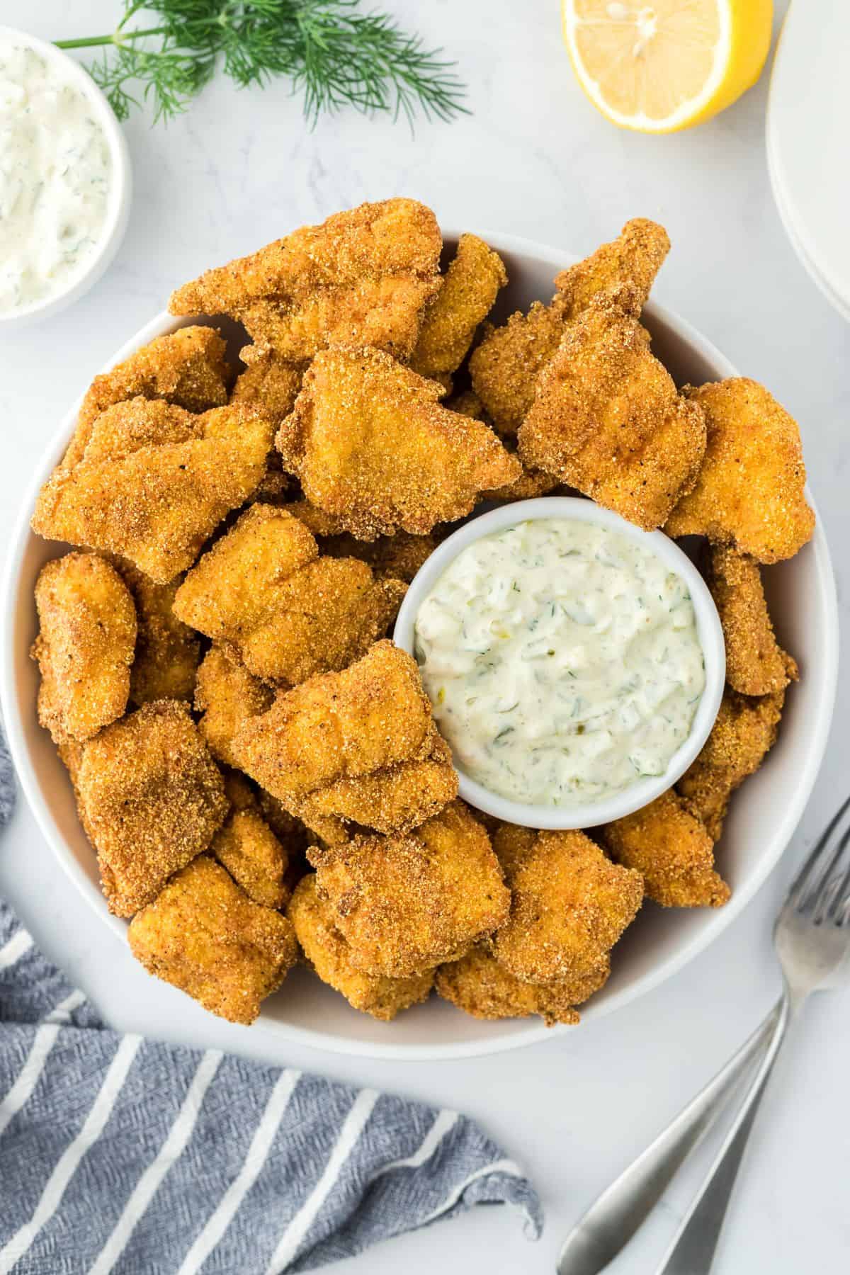 Overhead shot of catfish nuggets in a bowl with a side of tartar sauce, lemon, and dill on a white surface, with a grey and white striped cloth and forks next to it