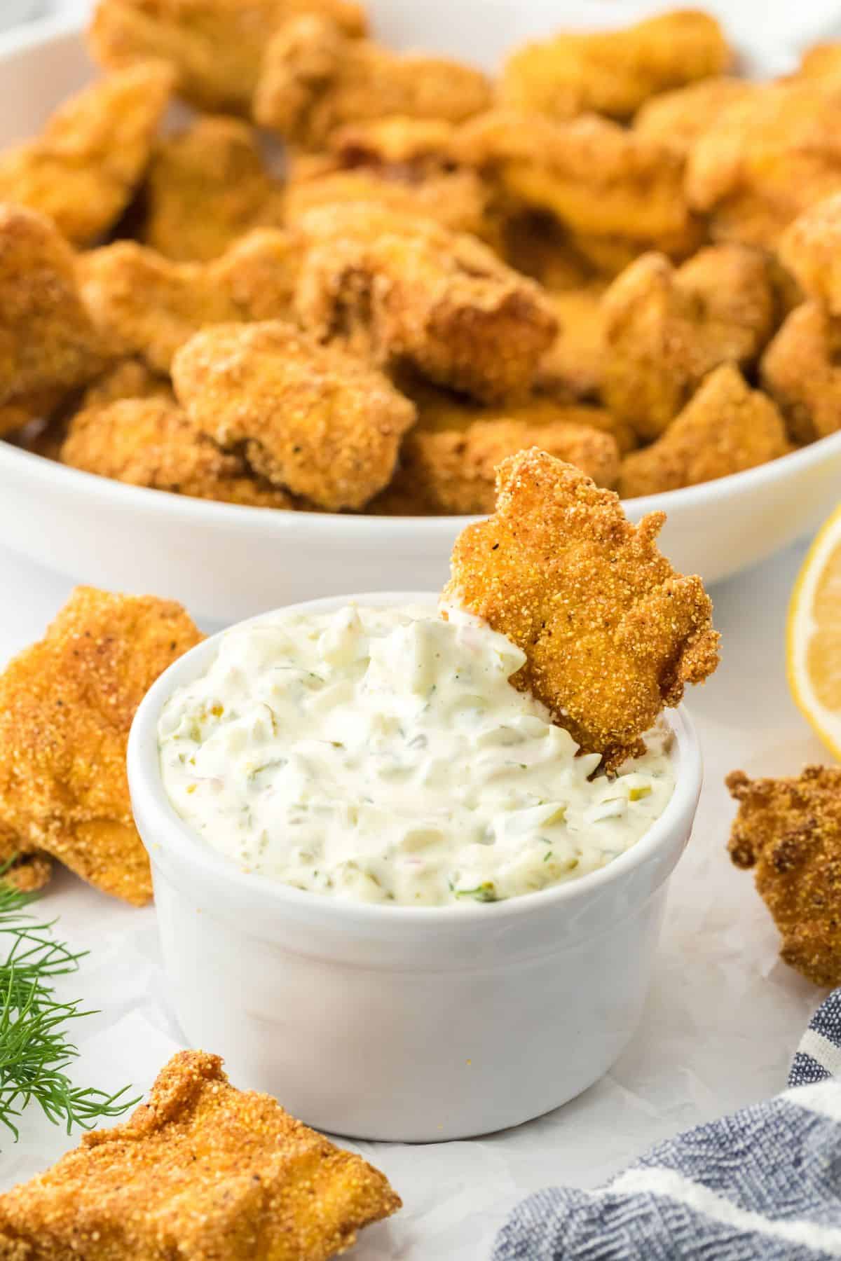 A catfish nugget dipped in a ramekin with tartar sauce, with more nuggets surrounding it and more nuggets in the background in a bowl