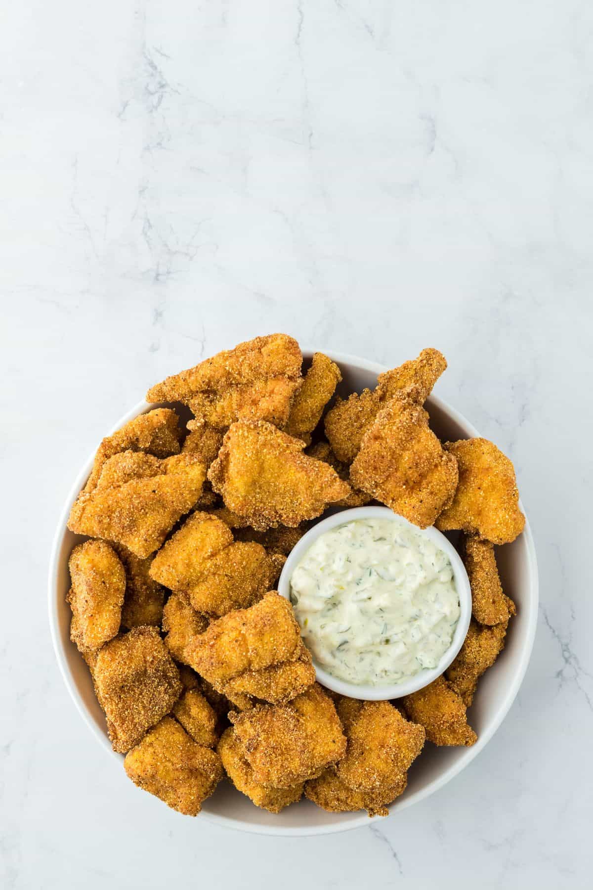 Overhead shot of a large bowl of catfish nuggets with a ramekin of tartar sauce in the center, set on a white marble surface