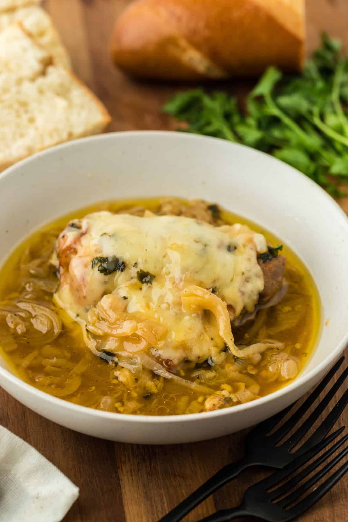French onion chicken in a white bowl, topped with melted cheese and a generous amount of caramelized onion broth. In the background, there's a baguette, fresh parsley, and two black forks