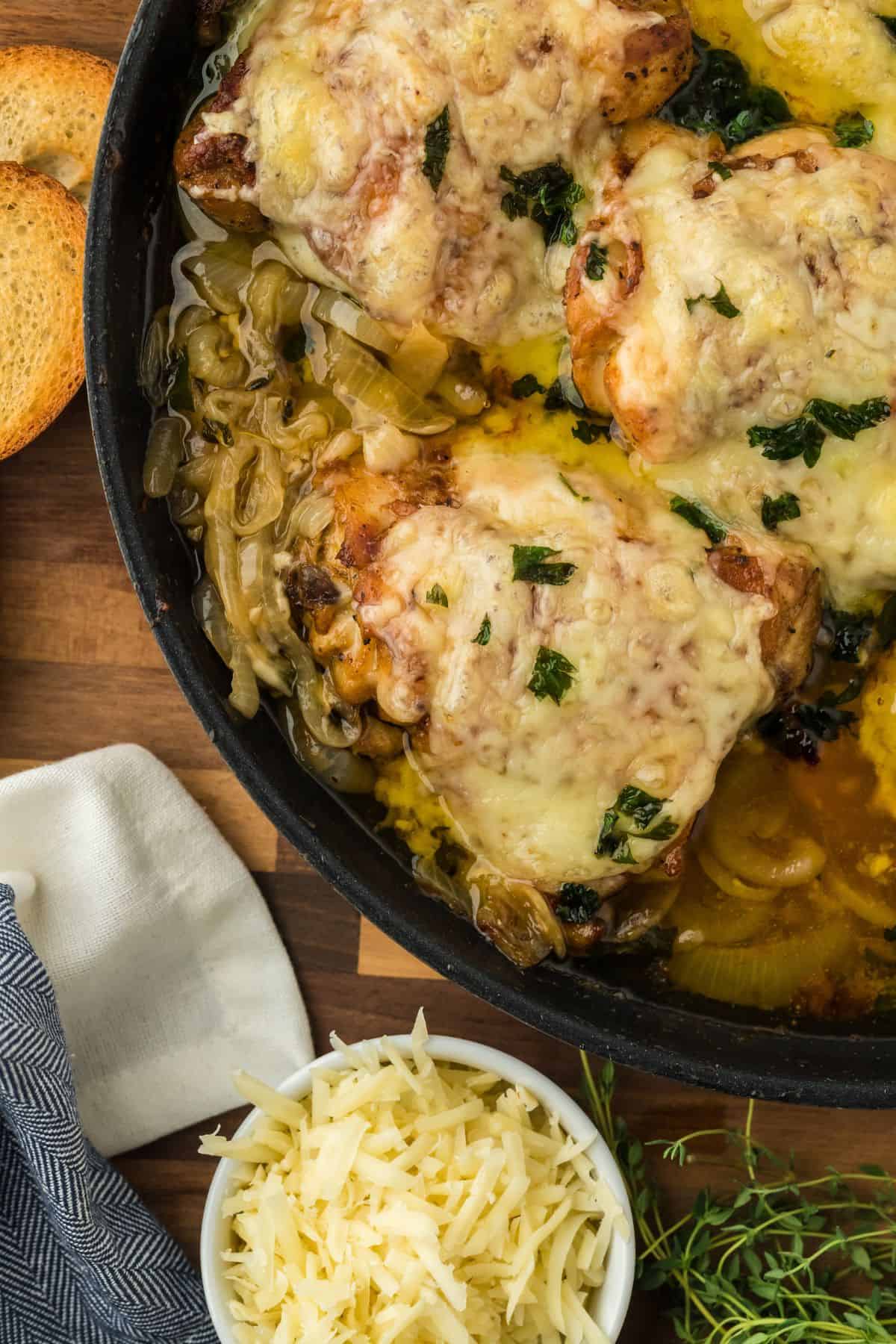 Closeup of french onion chicken in a skillet on a wooden cutting board. Next to it there are ingredients like shredded cheese, and toasted baguette slices