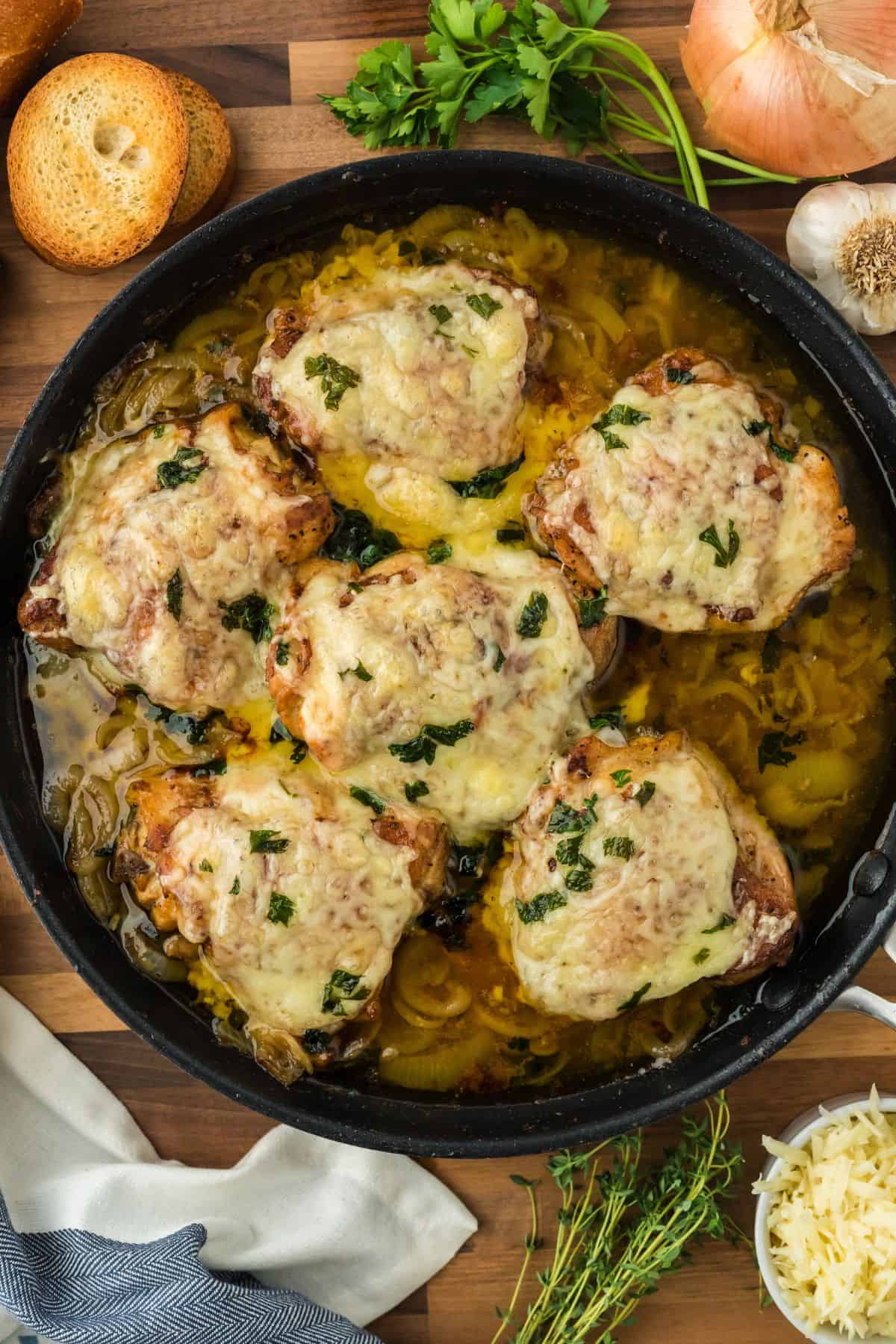 Overhead shot of french onion chicken in a skillet on a wooden cutting board. In the background, there are ingredients like onions, garlic, fresh parsley, a bunch of fresh thyme, shredded cheese, and toasted baguette slices