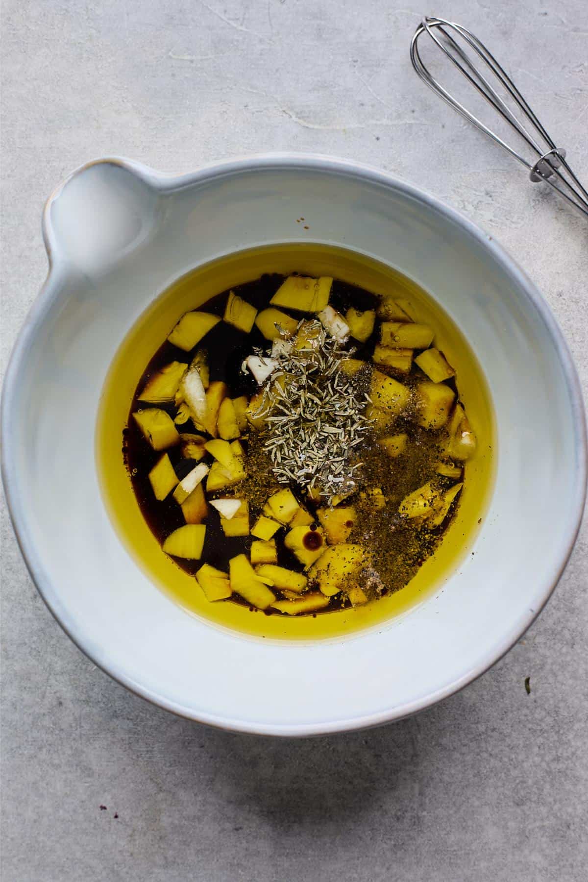 A white bowl containing oil, vinegar, garlic, herbs, and spices before whisking