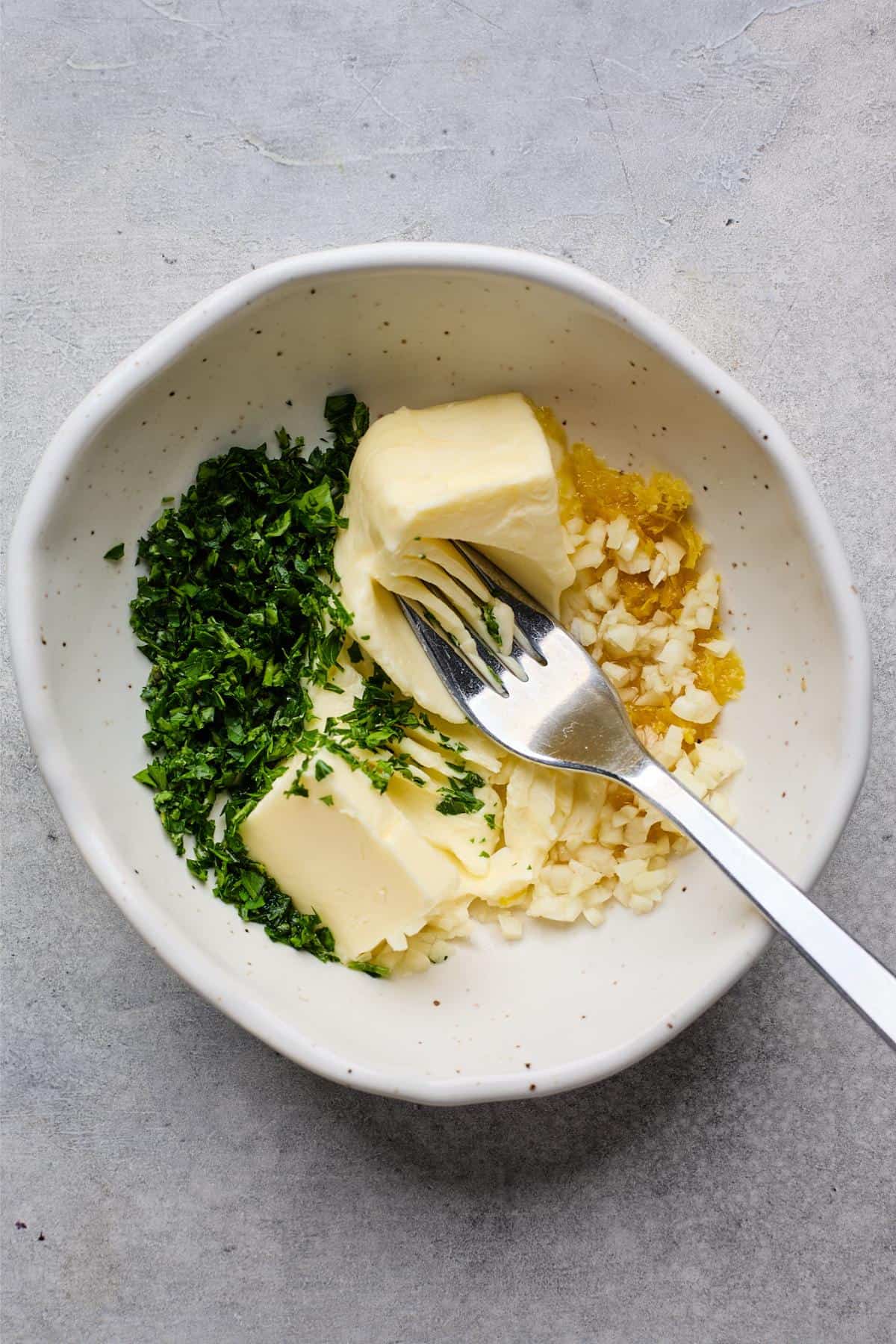 A small bowl with softened butter, chopped parsley, minced garlic, and lemon zest, with a fork