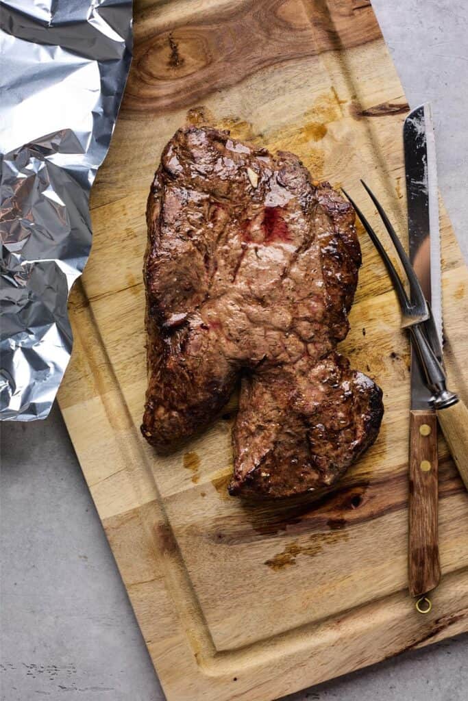 A cooked steak resting on a wooden cutting board with a carving fork and knife nearby