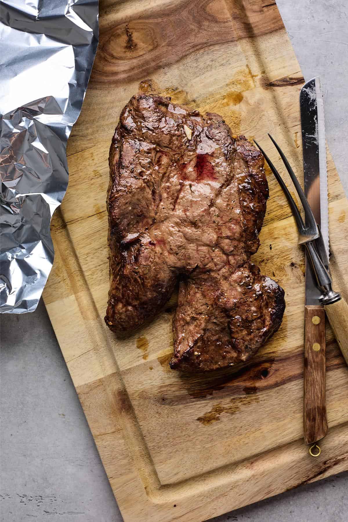 A cooked steak resting on a wooden cutting board with a carving fork and knife nearby