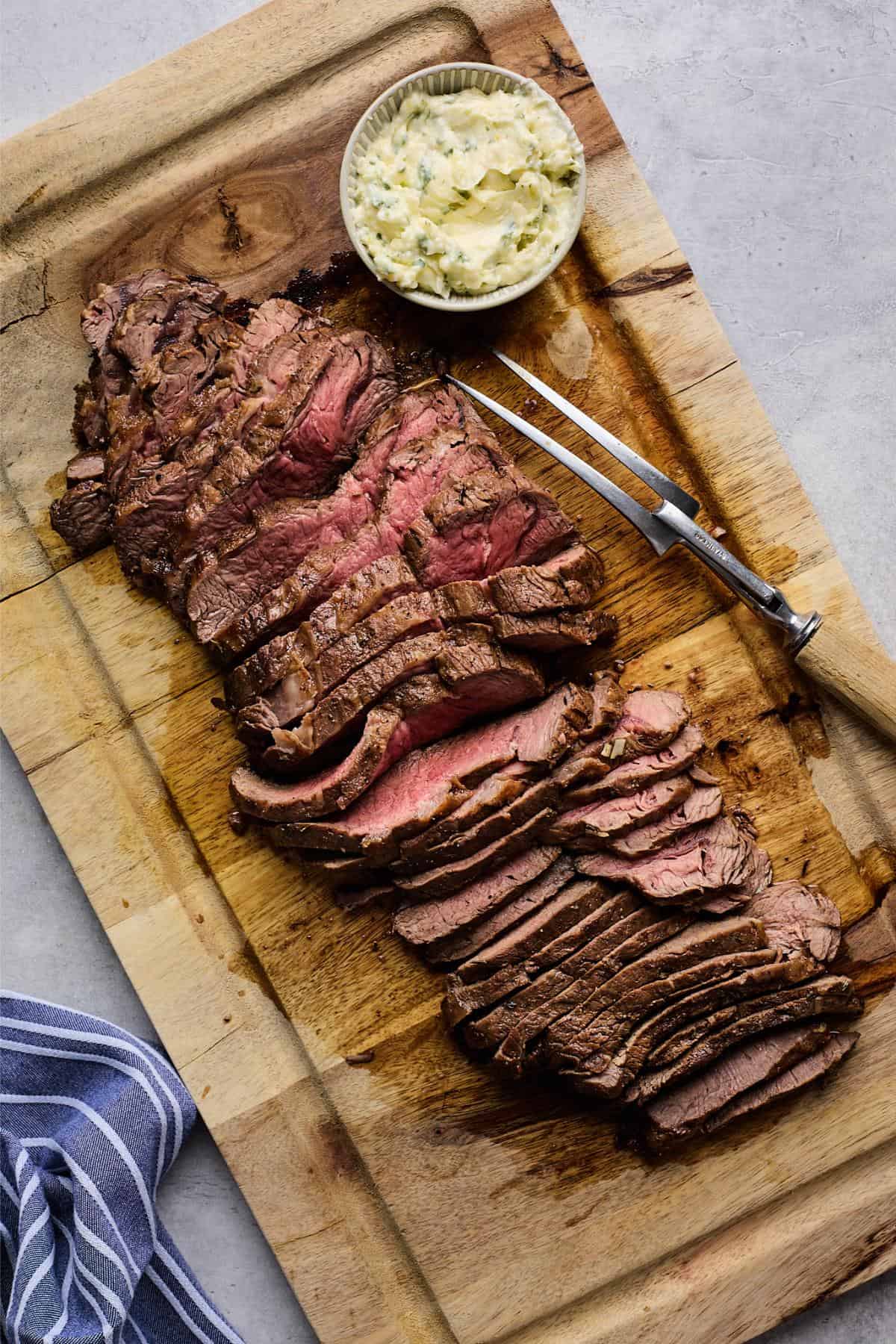 A sliced London broil arranged on a cutting board, showing a pink center, with a bowl of herb butter