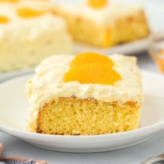 A slice of pig pickin' cake on a white plate, highlighting its moist yellow cake base and creamy frosting topped with mandarin orange slices. In the background, there's more cake and a bowl of mandarin oranges