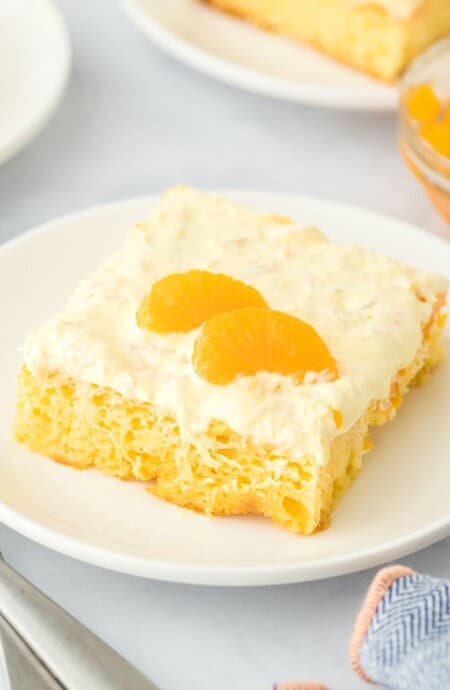 A slice of pig pickin' cake with a creamy frosting and mandarin oranges, served on a white plate. The background shows more slices of the cake on additional plates, with a bowl of mandarin oranges and forks