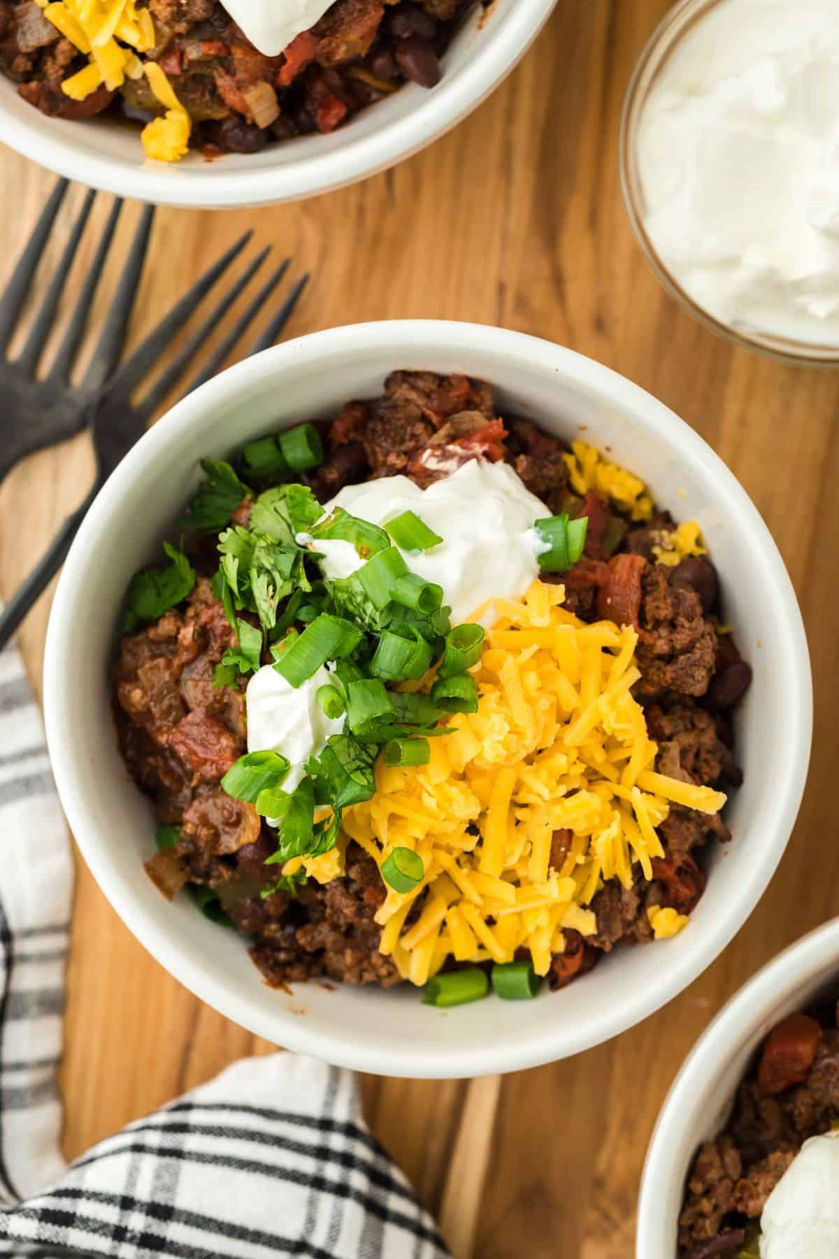 Overhead shot of a bowl of chili garnished with shredded cheese, sour cream and chopped green onions, with two forks, a small bowl of sour cream and more bowls of chili next to it