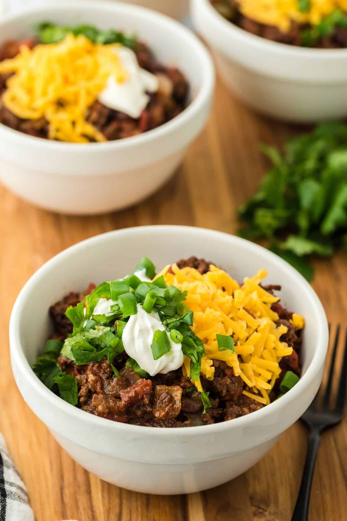 Sheet pan chili in a white bowl topped with shredded cheese, sour cream, and chopped green onions, with fresh cilantro and two more bowls of chili in the background
