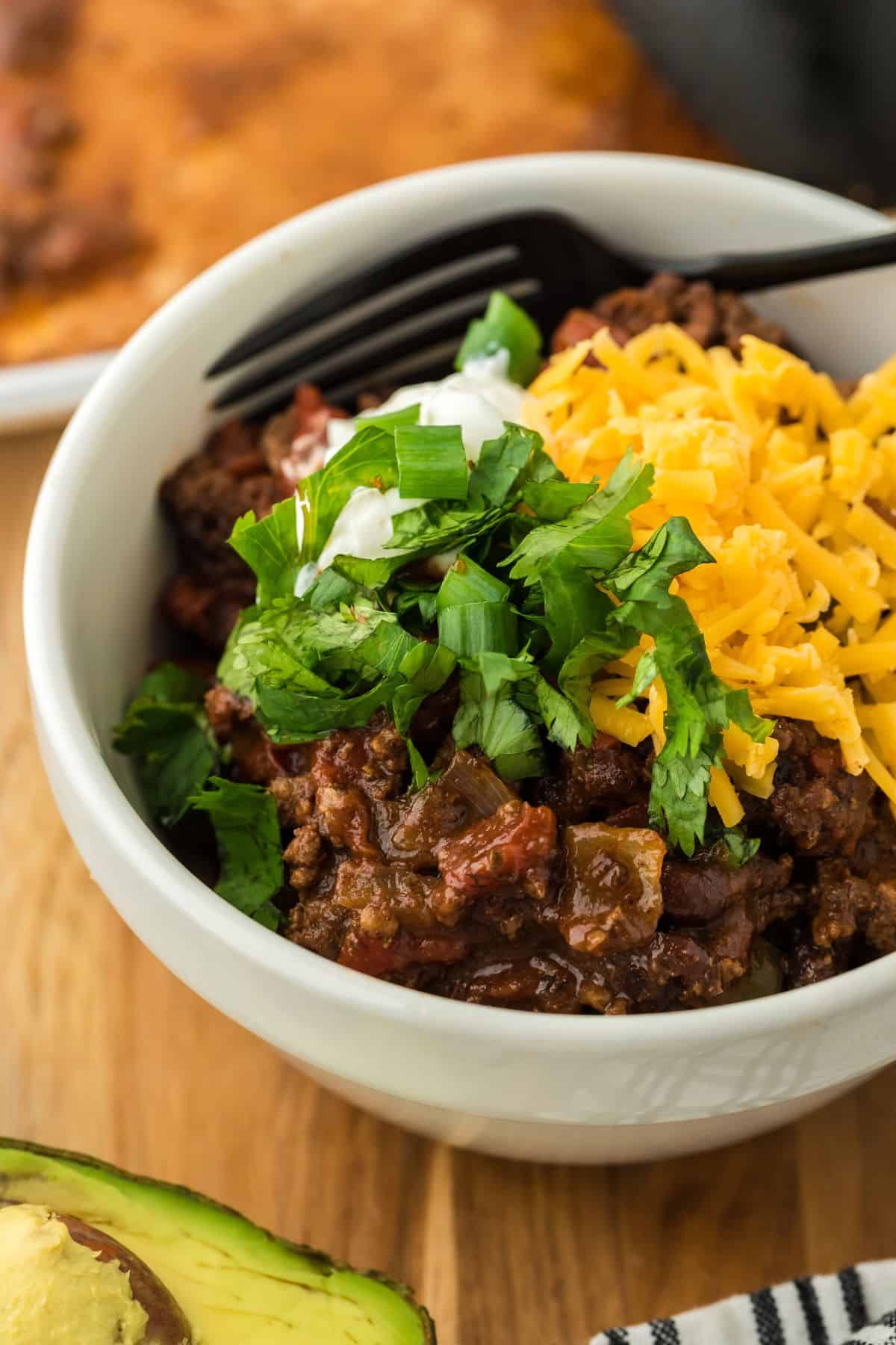 Closeup of a white bowl of sheet pan chili with a fork in it, topped with shredded cheese, a dollop of sour cream and fresh cilantro, placed on a wooden surface