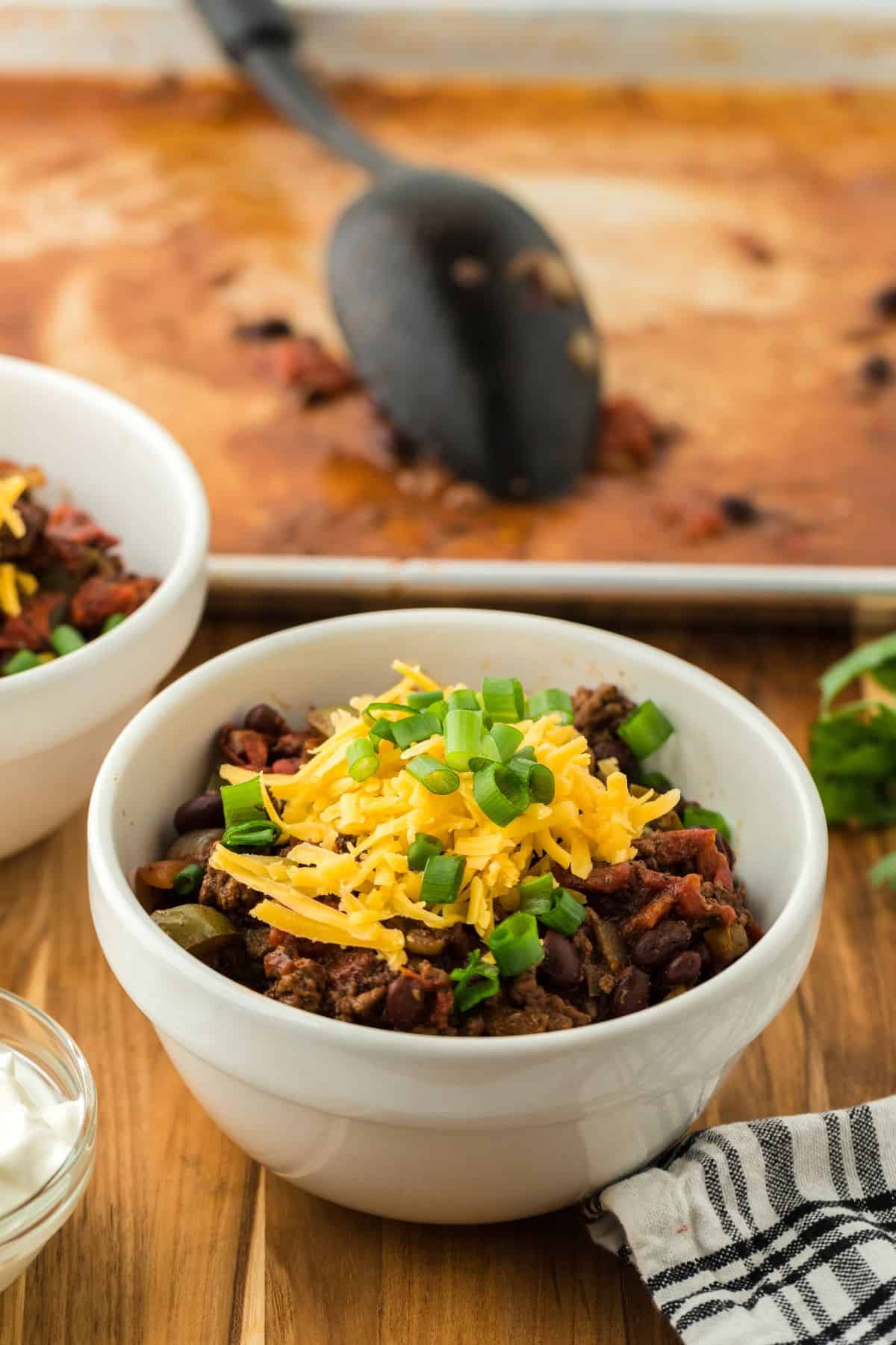 A bowl of sheet pan chili garnished with shredded cheese and green onions, with a black ladle in a sheet pan in the background