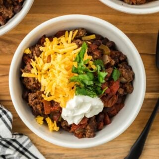 Overhead shot of a white bowl of sheet pan chili topped with shredded cheese and a dollop of sour cream, garnished with fresh cilantro, placed on a wooden surface with two black forks next to it