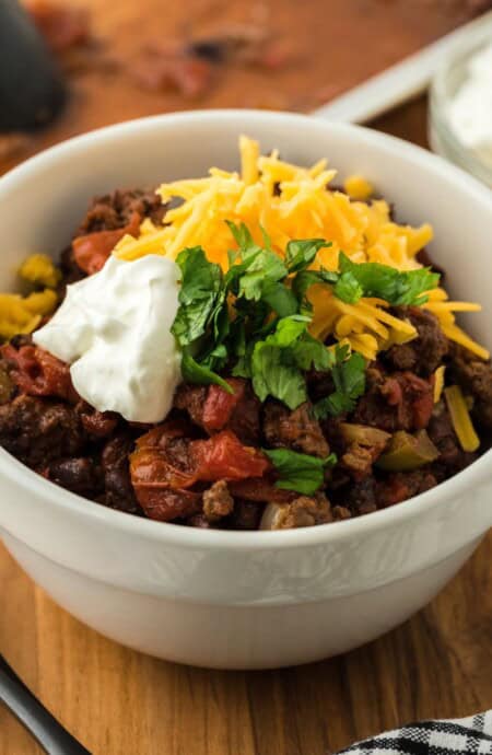 Sheet pan chili in a white bowl topped with shredded cheese, sour cream, and chopped green onions, with a bowl of sour cream and a sheet pan in the background