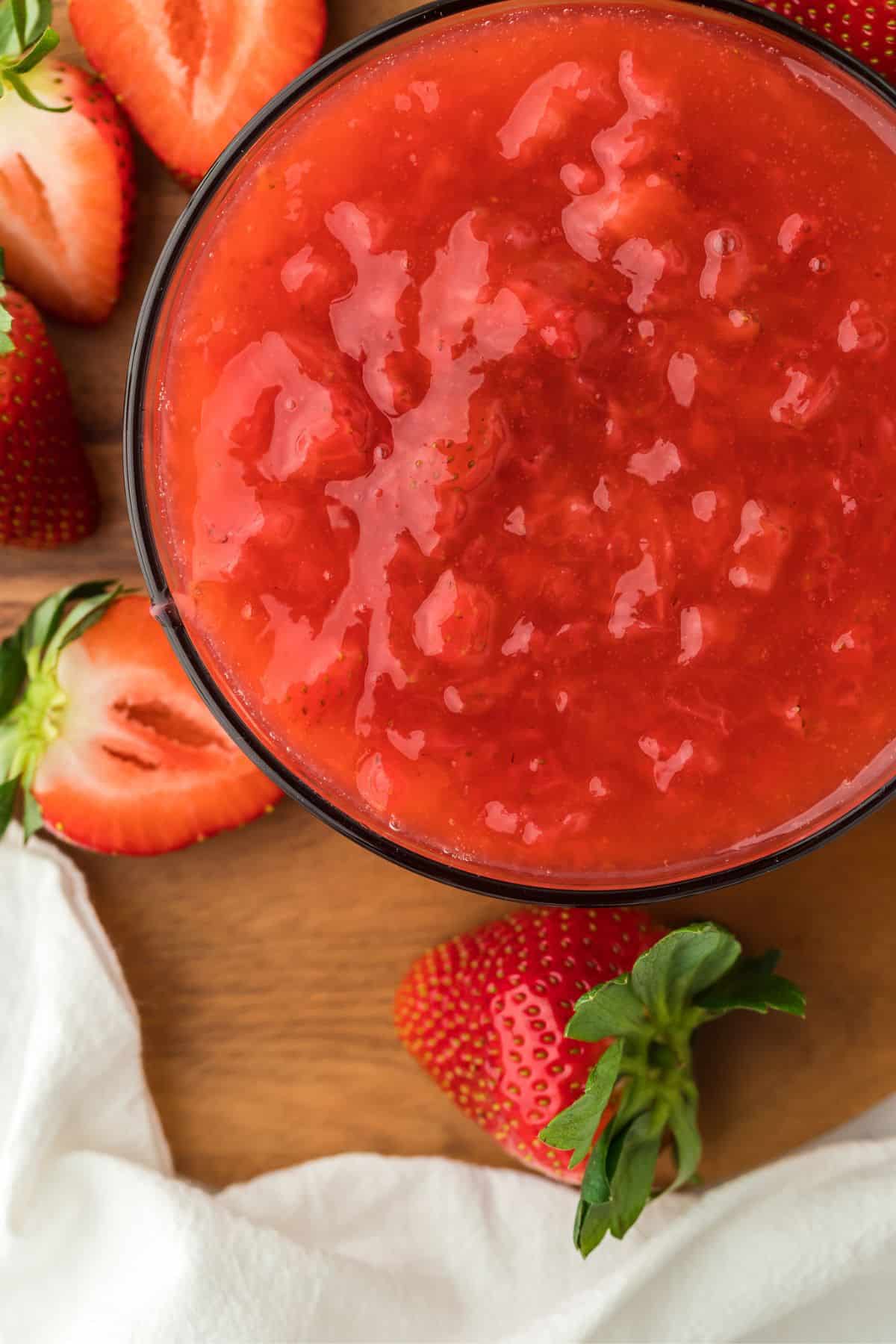 Closeup of a bowl of freshly made strawberry sauce in a wooden board. Surrounding the bowl are several strawberries and a white cloth