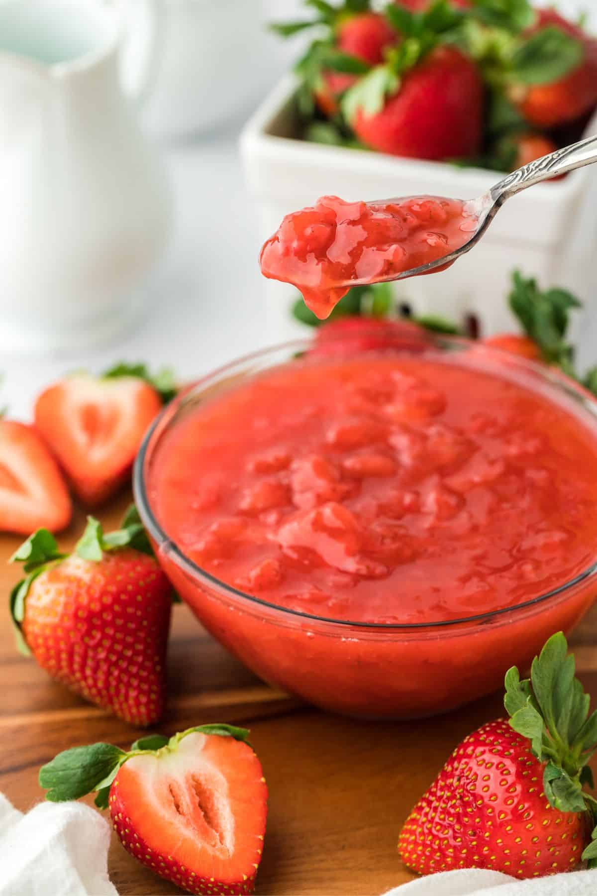 A spoon lifting a portion of strawberry sauce from a clear glass bowl. The bowl is surrounded by whole and halved fresh strawberries on a wooden surface. In the background, there's a white pitcher and a square white bowl with more strawberries