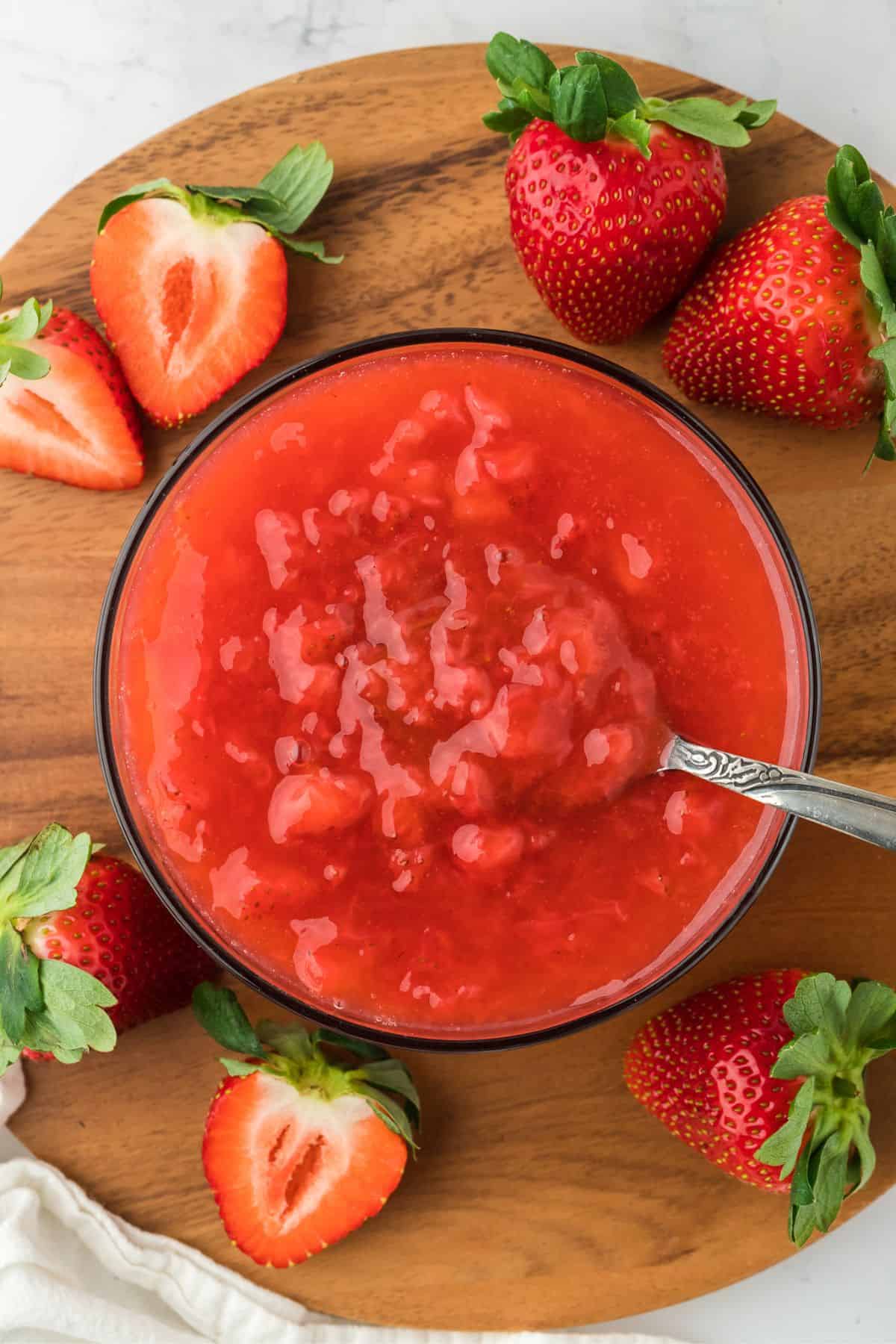 Overhead shot of a bowl of freshly made strawberry sauce in a wooden board with a spoon on it. Surrounding the bowl are several strawberries