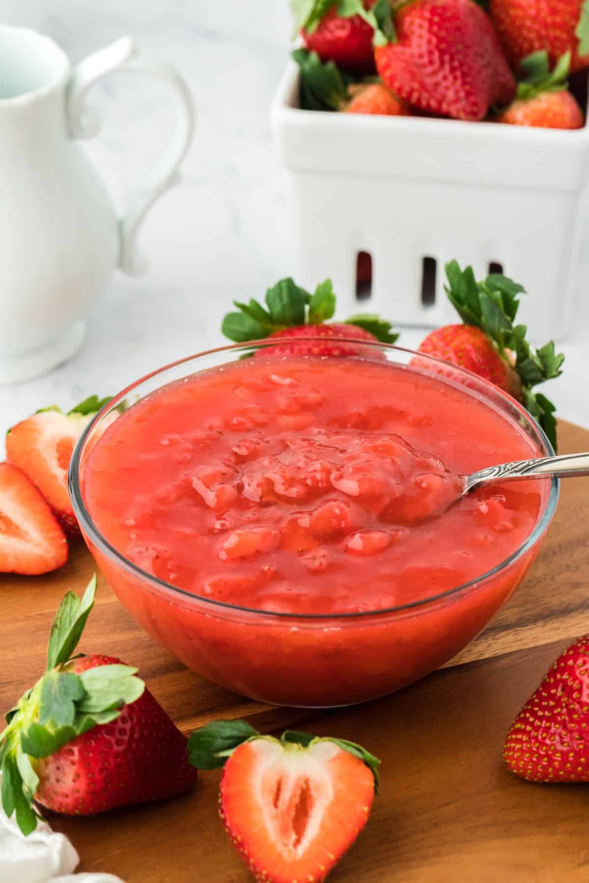 Strawberry sauce in a clear glass bowl with a spoon in it. The bowl is surrounded by whole and halved fresh strawberries on a wooden surface. In the background, there's a white pitcher and a square white bowl with more strawberries