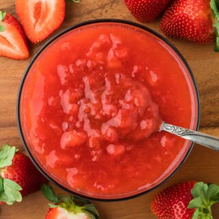 Overhead shot of a bowl of freshly made strawberry sauce with a spoon resting in the bowl. Surrounding the bowl are several strawberries
