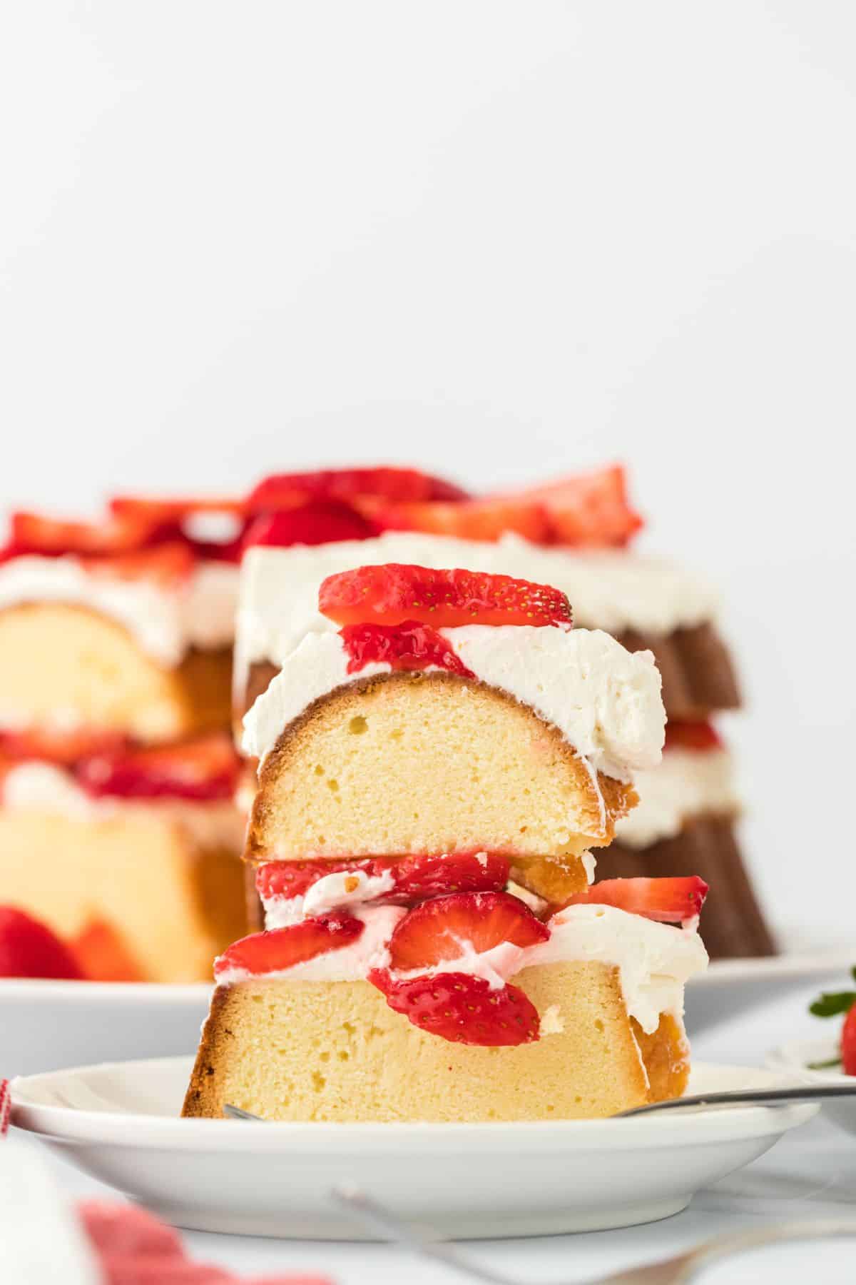 Closeup of a slice of strawberry shortcake pound cake on a plate, showing the texture of the cake and the layers of whipped cream and fresh strawberry slices, with more of the cake in the background