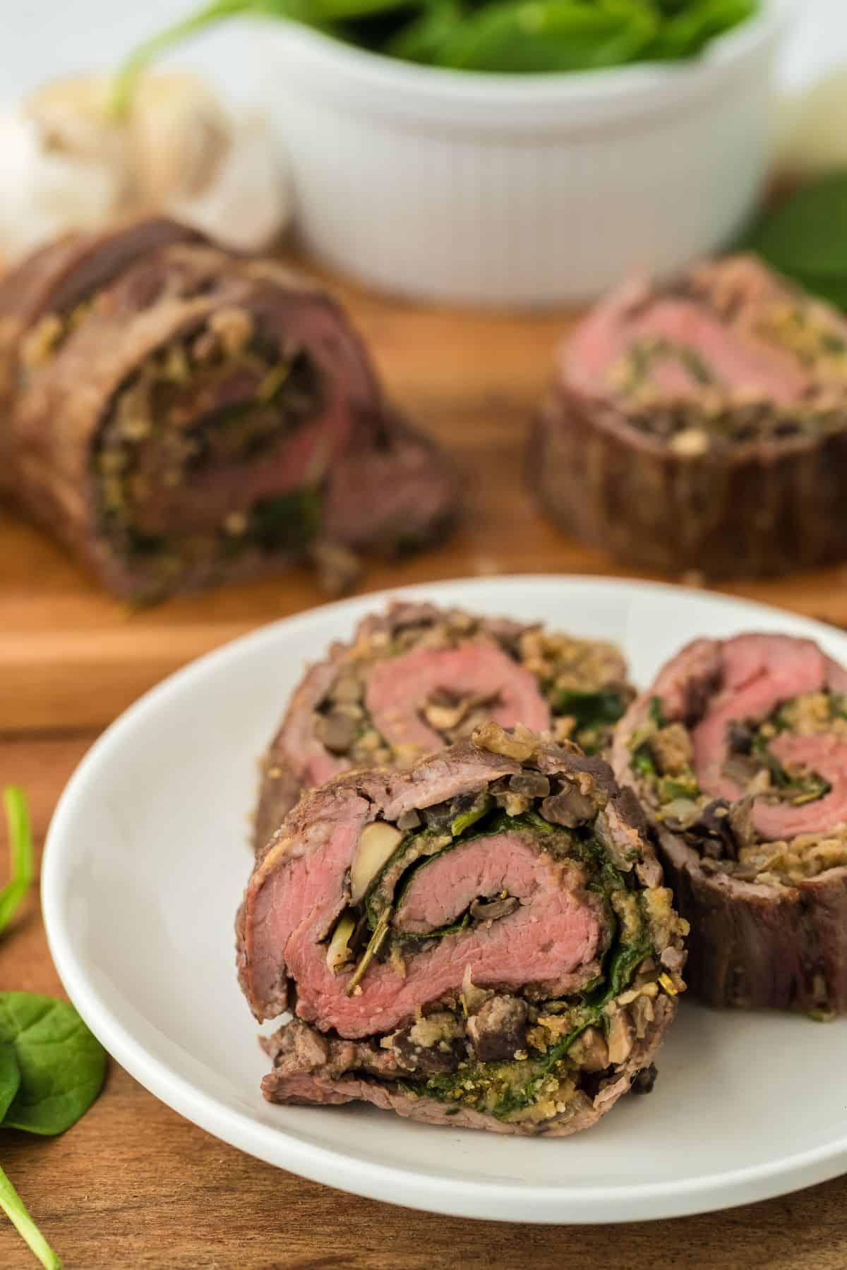 Closeup of slices of stuffed flank steak filled with a spinach and mushroom mixture on a white plate, with more steak rolls in the background