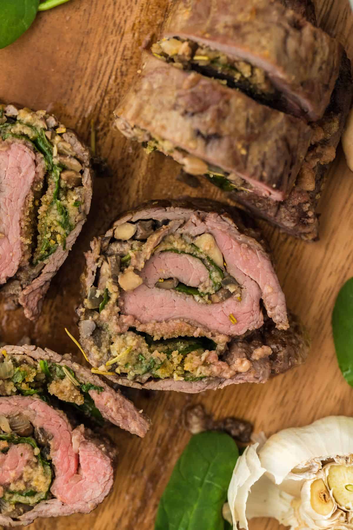 Closeup of several sliced stuffed flank steak with spinach and mushroom stuffing on a wooden cutting board. There are fresh spinach leaves and a head of garlic next to it