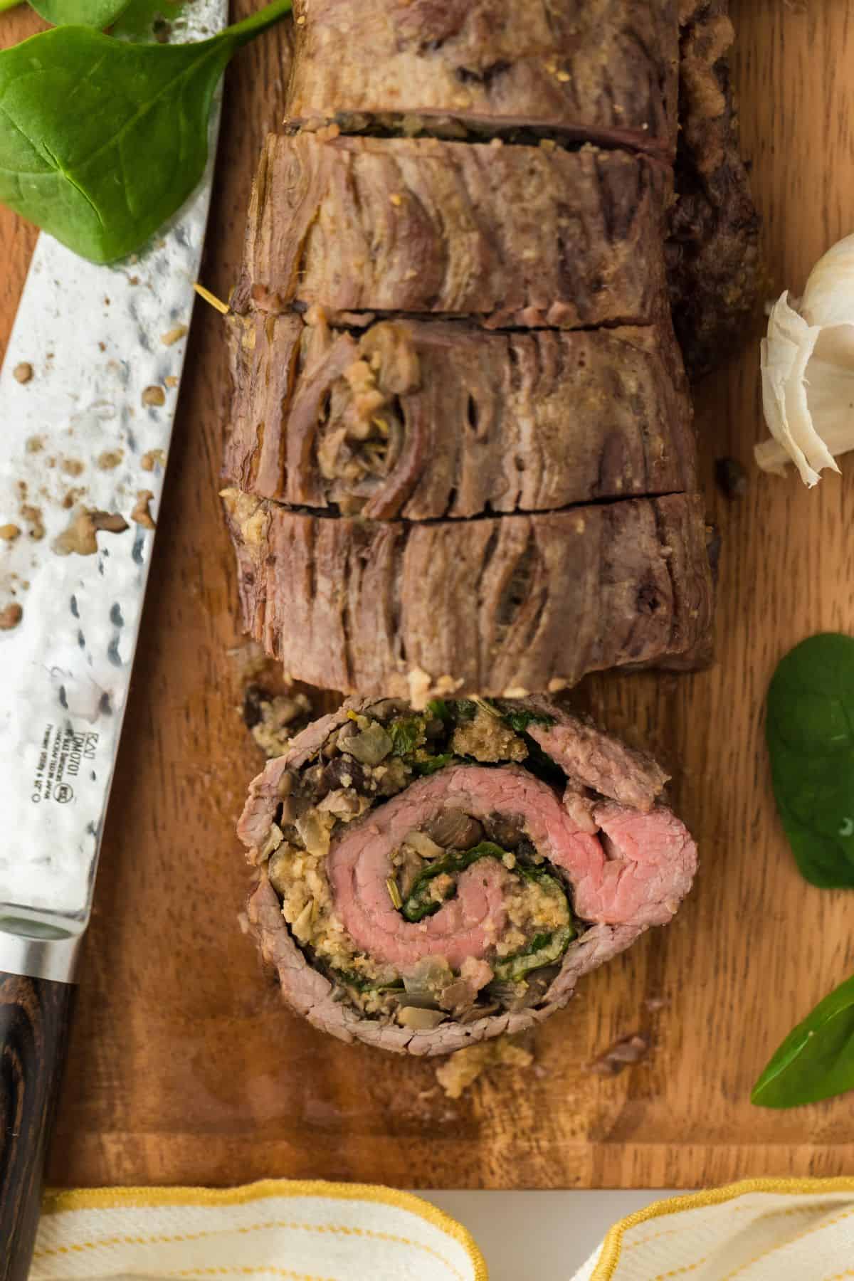 Overhead shot of a stuffed flank steak with mushroom and spinach filling, cut into slices on a wooden cutting board, with a knife and fresh spinach next to it