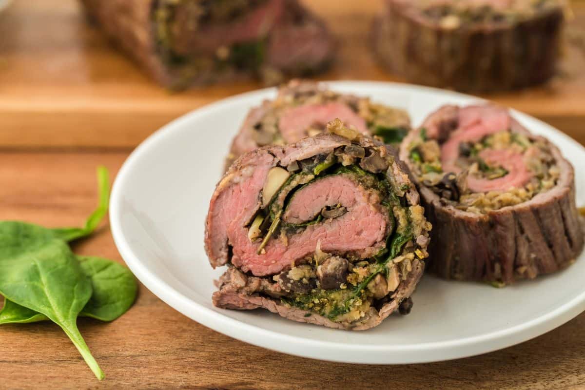Slices of stuffed flank steak on a white plate, with fresh spinach leaves next to it and more rolls in the background