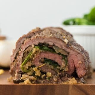 Closeup of a stuffed flank steak with a green spinach and mushroom filling, presented on a wooden cutting board with fresh greens in the background