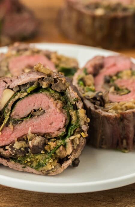 Closeup of slices of stuffed flank steak filled with a spinach and mushroom mixture on a white plate, with another steak roll in the background