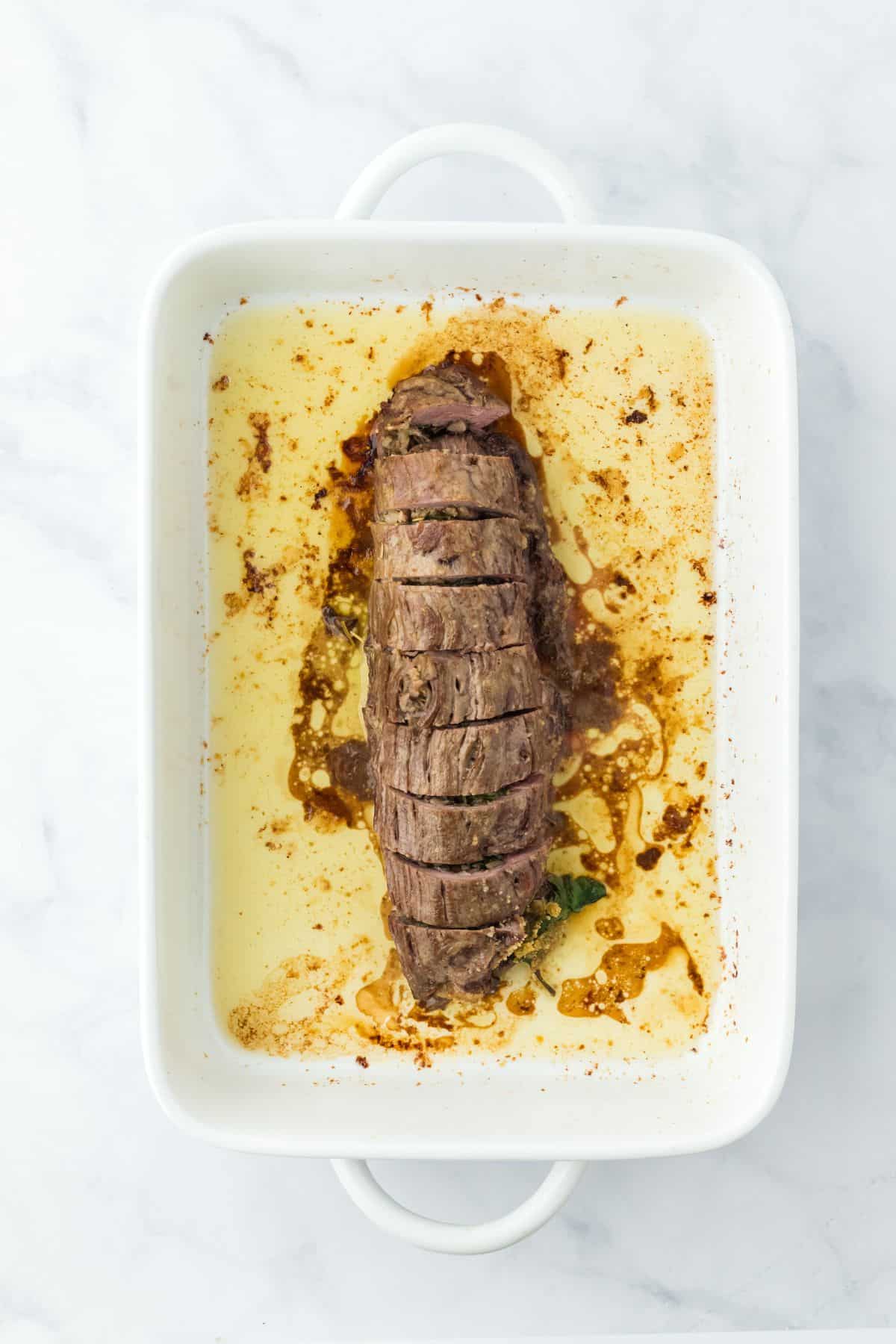 A whole stuffed flank steak sliced and placed in a white baking dish, with cooking juices around it, set on a marble countertop