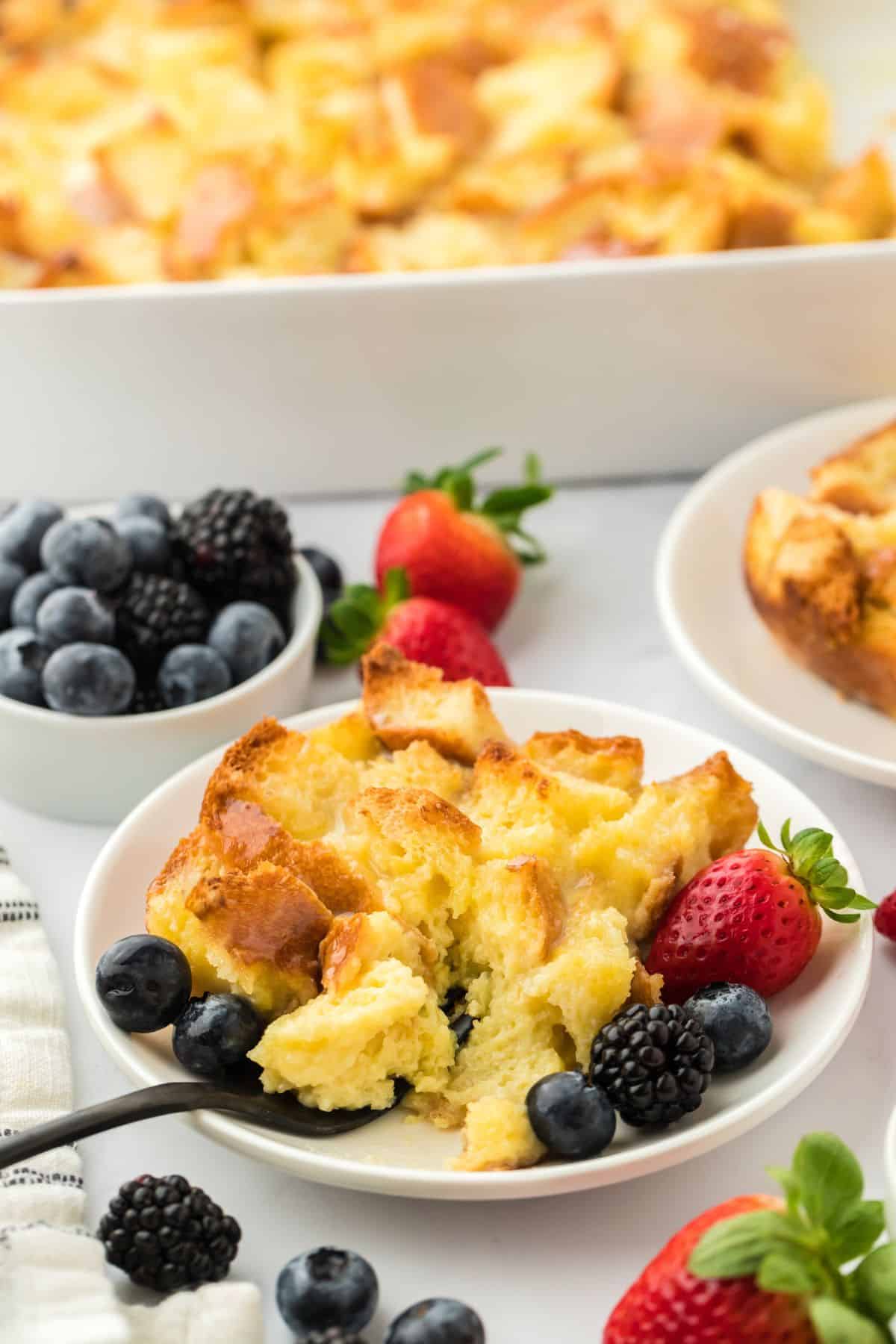 A white plate with a serving of white chocolate bread pudding with a black fork and berries next to it. In the background, there's a baking dish with more bread pudding, and a small bowl filled with an assortment of fresh berries like strawberries, blueberries, and blackberries
