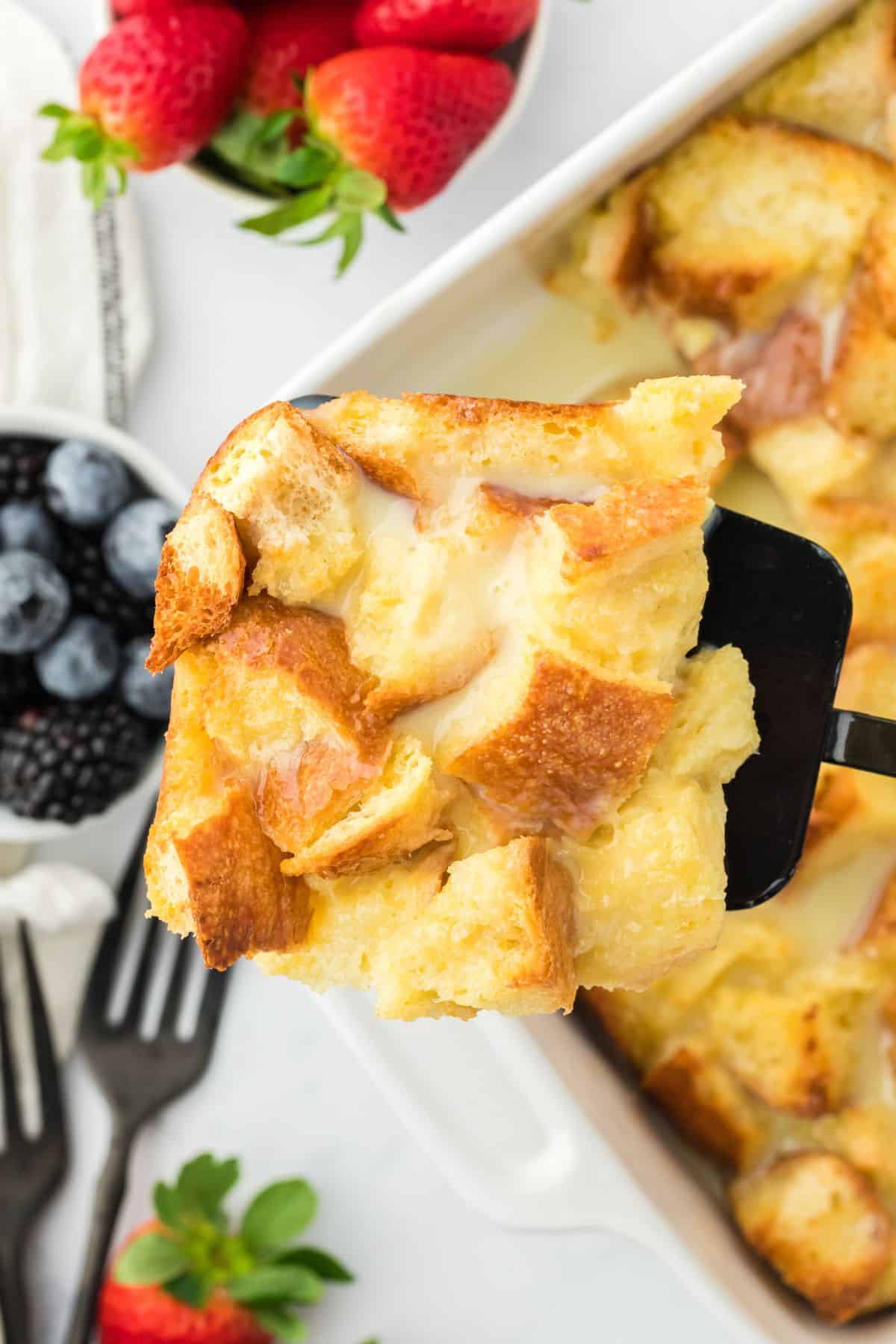 Closeup of a serving spoon lifting a generous piece of white chocolate bread pudding out of a white baking dish. In the background, there is a bowl of mixed berries and black forks