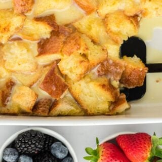 Closeup of white chocolate bread pudding in a white baking dish, with a serving spoon lifting a portion. Next to it there are small bowls of blackberries, blueberries and strawberries