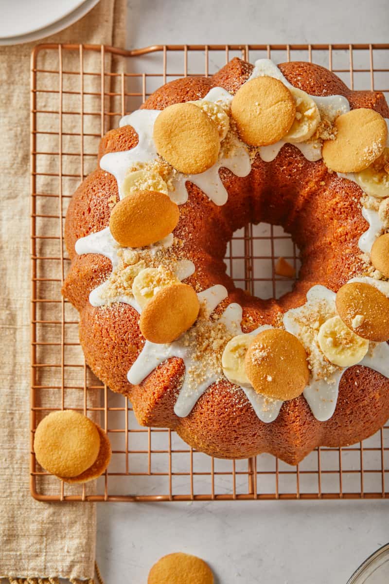 Overhead shot of banana pudding pound cake with white glaze, topped with banana slices and vanilla wafers, sitting on a copper cooling rack, with more wafers scattered around