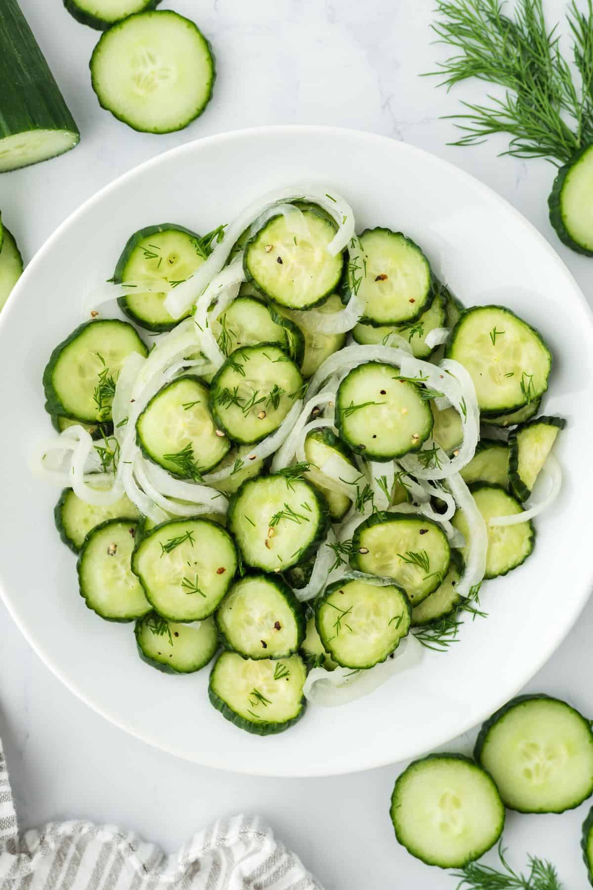 Overhead shot of cucumber and onion salad garnished with dill, arranged in a white plate. Next to it there are more cucumber slices and sprigs of dill