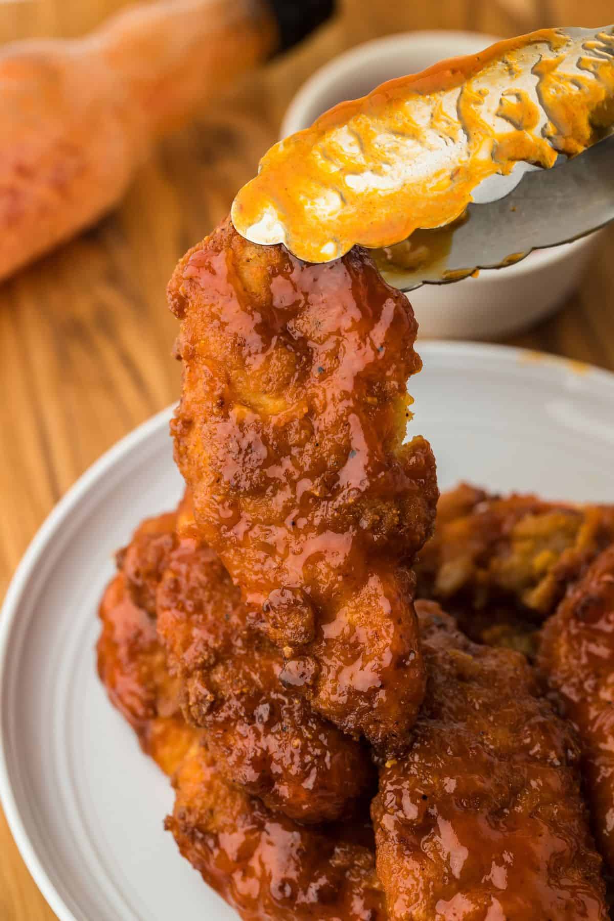 Tongs holding a hot honey chicken tender over a plate stacked with more glazed tenders, all on a wooden table