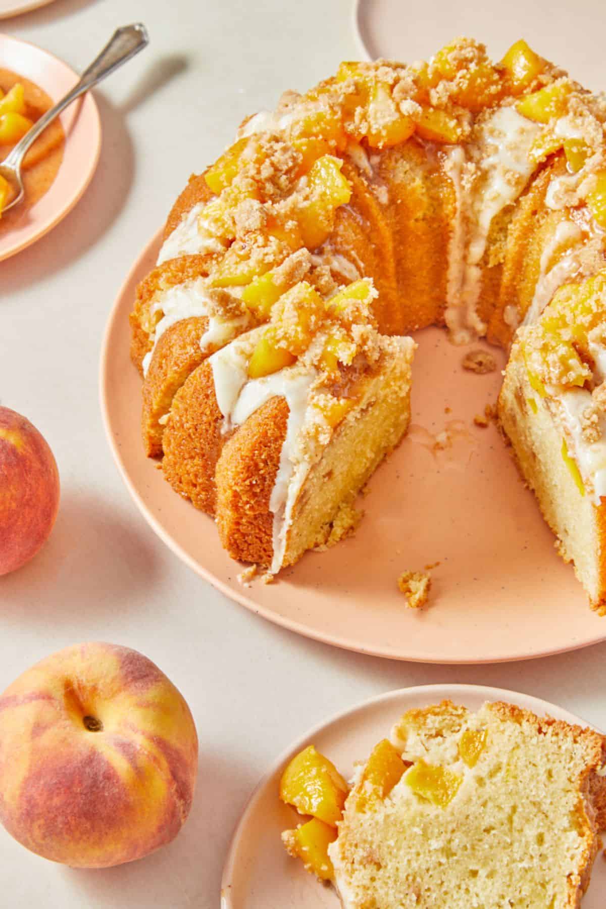 A sliced peach cobbler pound cake with one piece removed, showing the cake’s interior. The cake is garnished with peach chunks and crumble topping, drizzled with glaze, and accompanied by whole peaches, all set on a pink plate