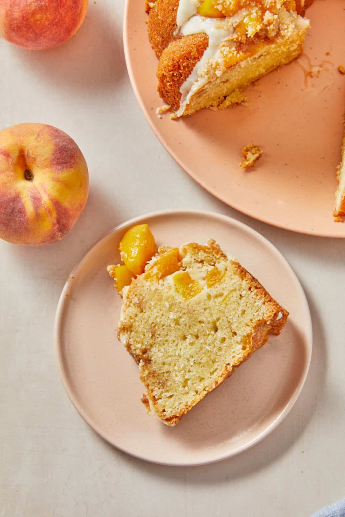 A slice of peach cobbler pound cake on a pink plate, showing its moist texture and peach pieces on top, with a whole peach beside it and a plate with more cake behind it