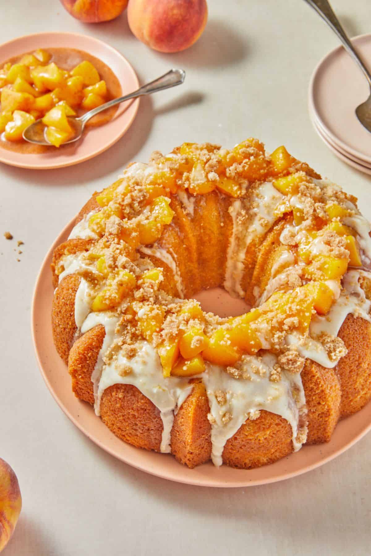 A peach cobbler pound cake drizzled with white glaze and topped with fresh peach pieces and crumble, served on a pink plate, with empty plates, peaches, and a plate with more peach pieces in the background