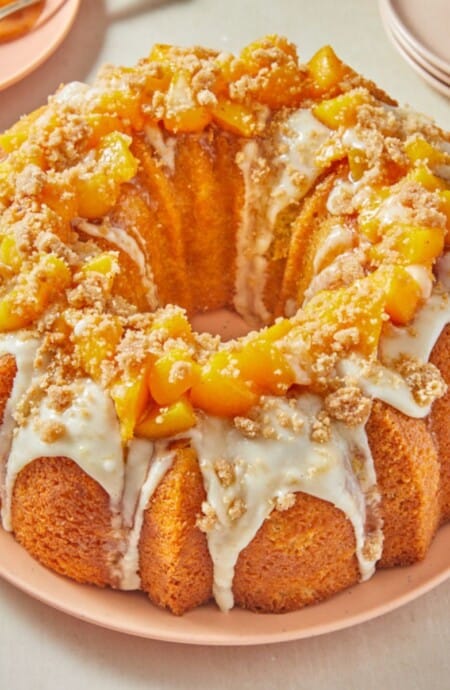 A peach cobbler pound cake drizzled with white glaze and topped with fresh peach pieces and crumble, served on a pink plate